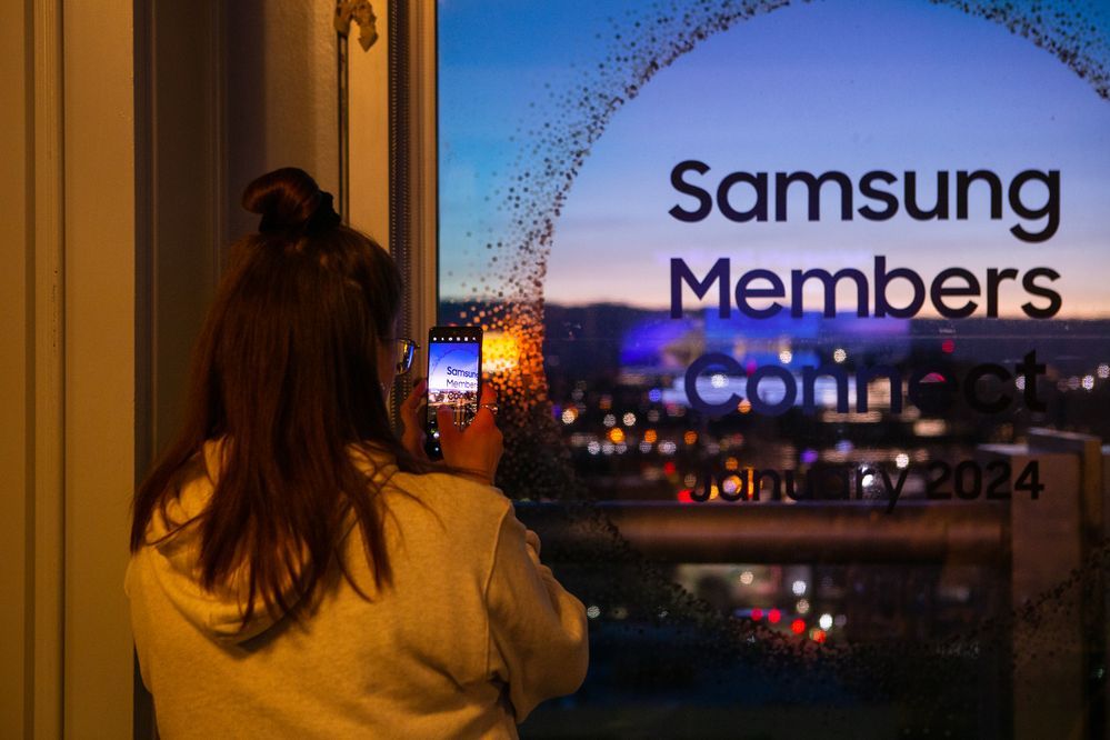 A woman taking a photo of the evening skyline with Samsung Members Connect written on the window.jpg